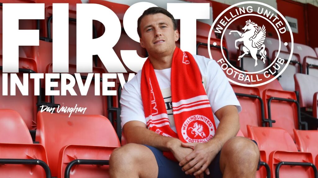 Dan Vaughan sits in the stadium seats wearing a red Welling United FC scarf during his first interview as a new signing for the club.